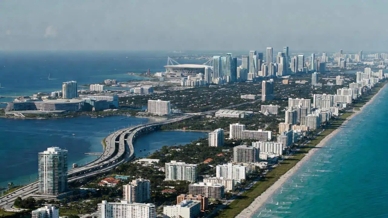Aerial view of Miami Beach's hotel-lined coastline and sandy beach along the Atlantic Ocean, with turquoise waters in the foreground. A curved highway spans Biscayne Bay, leading toward the dense Brickell and downtown Miami skyline in the distance. Hard Rock Stadium is faintly visible far off under a partly cloudy sky, capturing Miami's coastal-to-urban landscape.